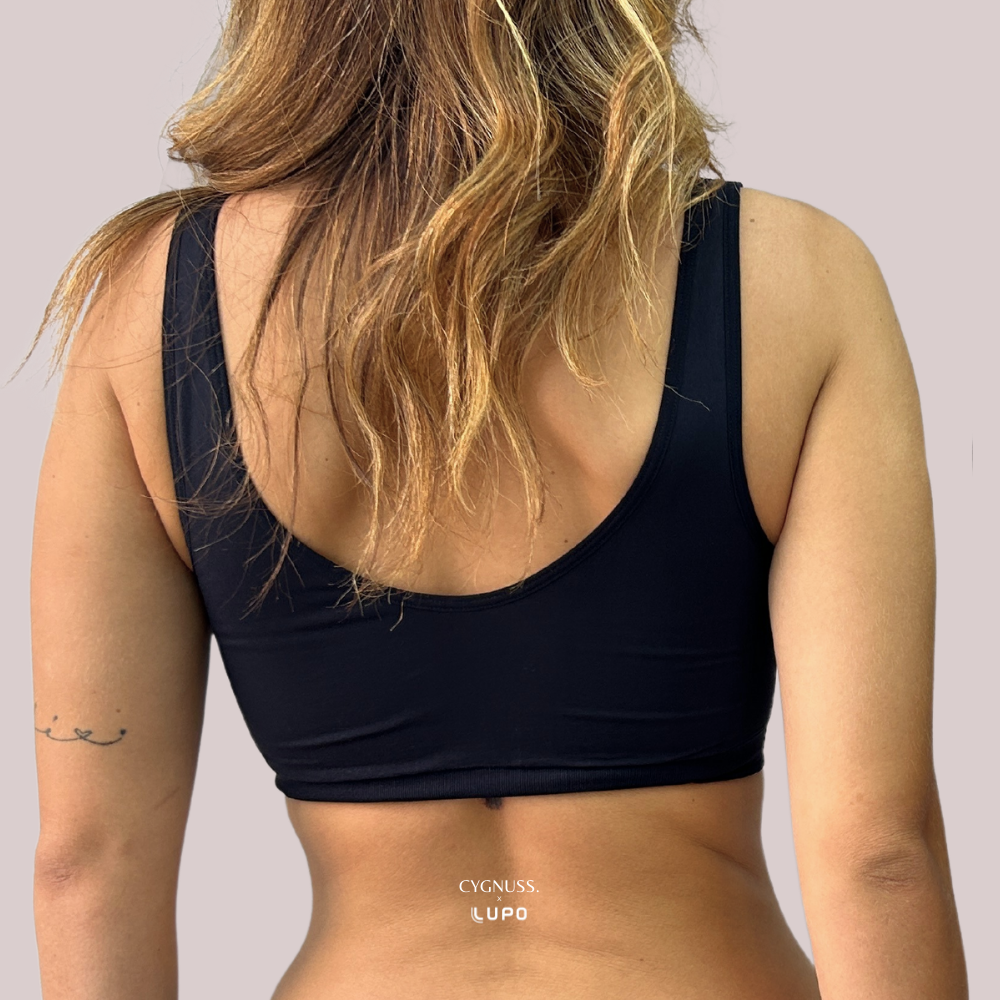 Imagem mostra a parte de trás de uma mulher usando top preto sem costura da marca Cygnuss em parceria com Lupo. O modelo tem alças largas e costas cavadas, proporcionando sustentação e conforto. O fundo neutro destaca a peça.