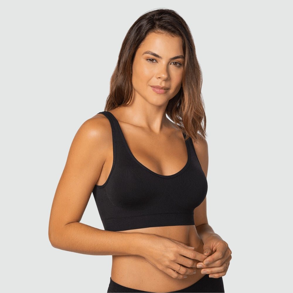 Mulher em pose frontal vestindo top preto básico sem costura da Cygnuss em parceria com a Lupo. A peça possui bojo anatômico, alças largas e cós reforçado, oferecendo sustentação com conforto. O fundo cinza claro contribui para destacar o design funcional e minimalista do top.
