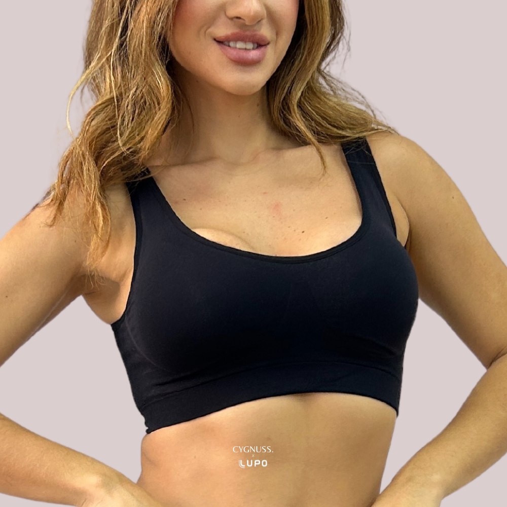 Mulher sorrindo com top preto sem costura da Cygnuss em parceria com Lupo. A peça possui bojo anatômico, alças largas e cós reforçado que proporciona sustentação e conforto. O fundo neutro destaca o design minimalista do top.