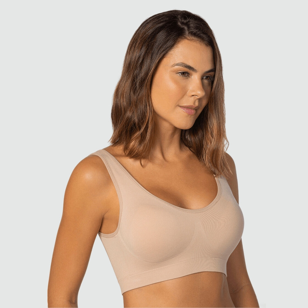 Mulher de perfil vestindo top nude sem costura da Cygnuss em parceria com a Lupo, com alças largas e bojo embutido. O tecido suave acompanha o corpo, oferecendo sustentação e um visual elegante. Fundo cinza claro realça os tons naturais da peça e da modelo.