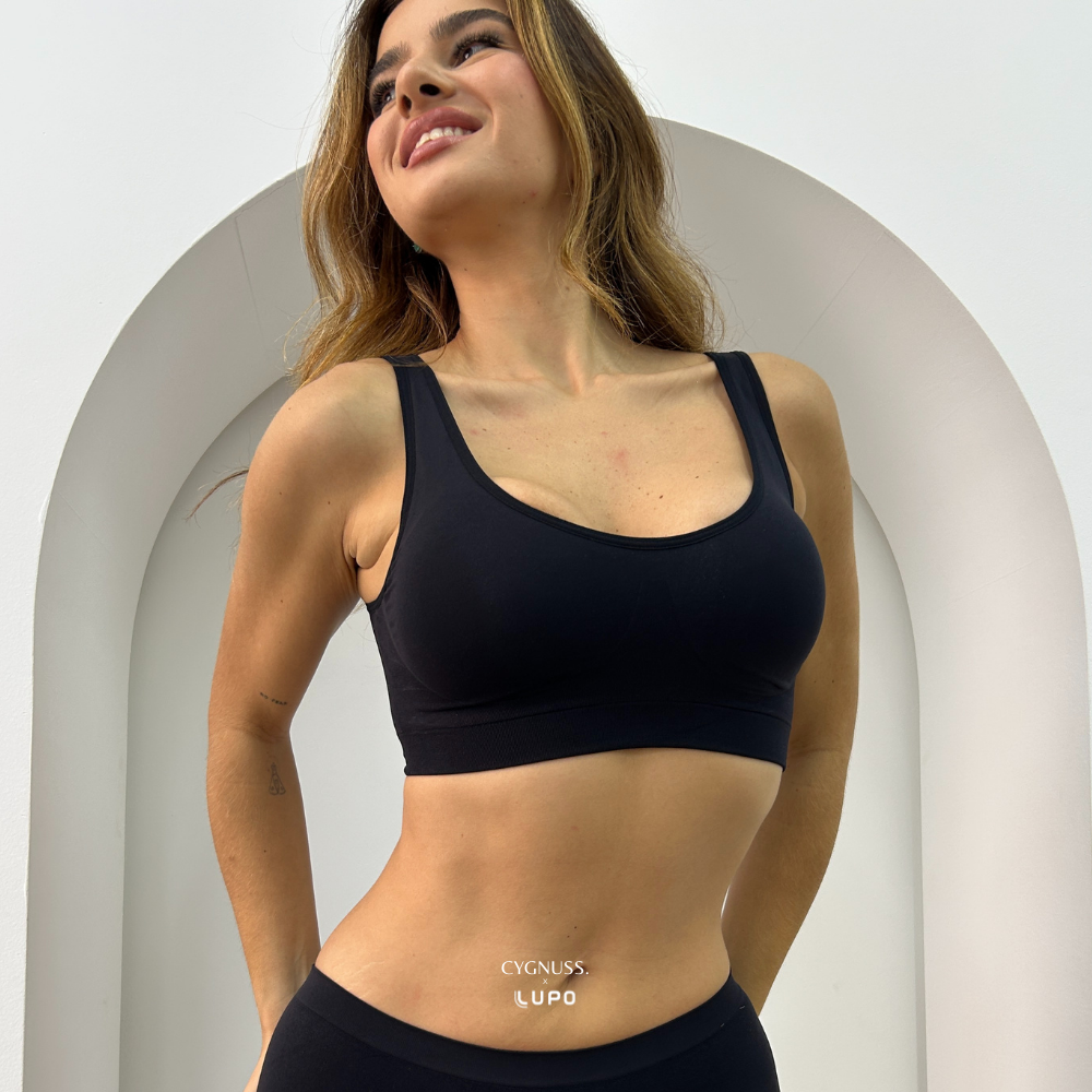 Mulher sorridente usando top preto estruturado da Cygnuss em parceria com a Lupo. A peça sem costura possui alças largas, bojo anatômico e cós reforçado. O fundo branco com arco moderno valoriza a elegância e o design clean da imagem.