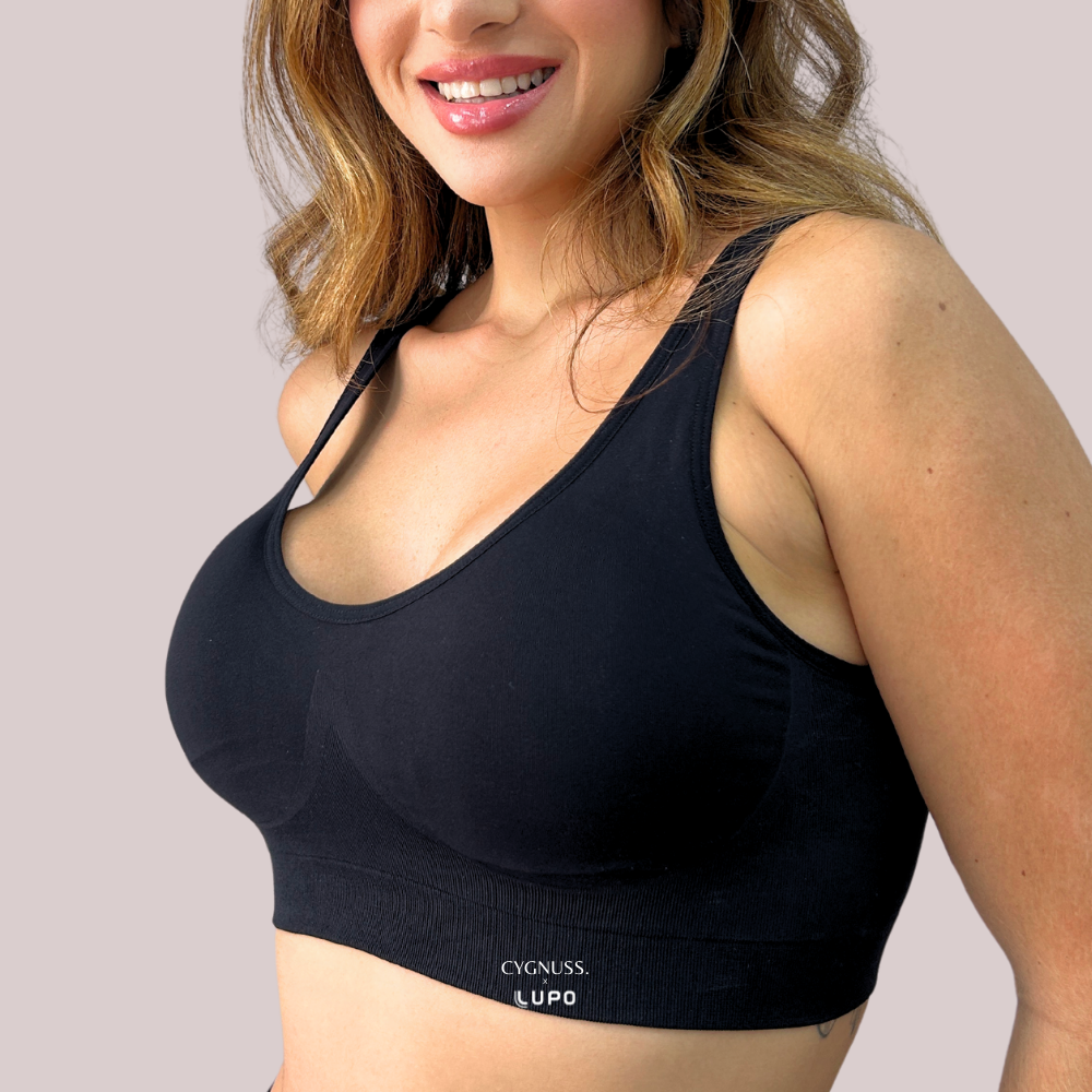 Mulher sorridente vestindo um top preto sem costura com bojo anatômico e alças largas. A peça modela suavemente o busto e tem as marcas “Cygnuss” e “Lupo” estampadas na base. O fundo é neutro, destacando o conforto e ajuste do top.