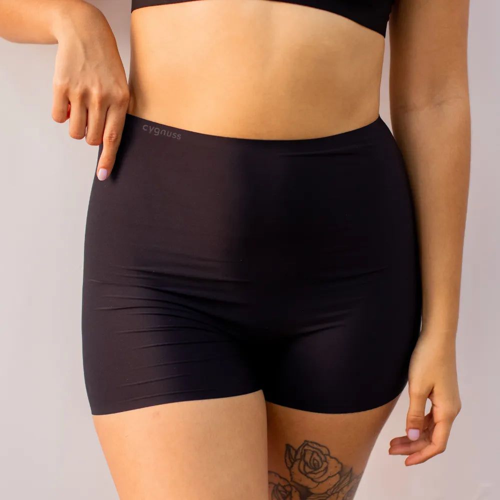 Imagem de uma mulher usando o Short Segunda Pele Sem Costura na cor preta. A peça proporciona conforto e modelagem sem costuras, garantindo um ajuste perfeito e discreto para o dia a dia, com o logo da Cygnuss visível na parte superior.