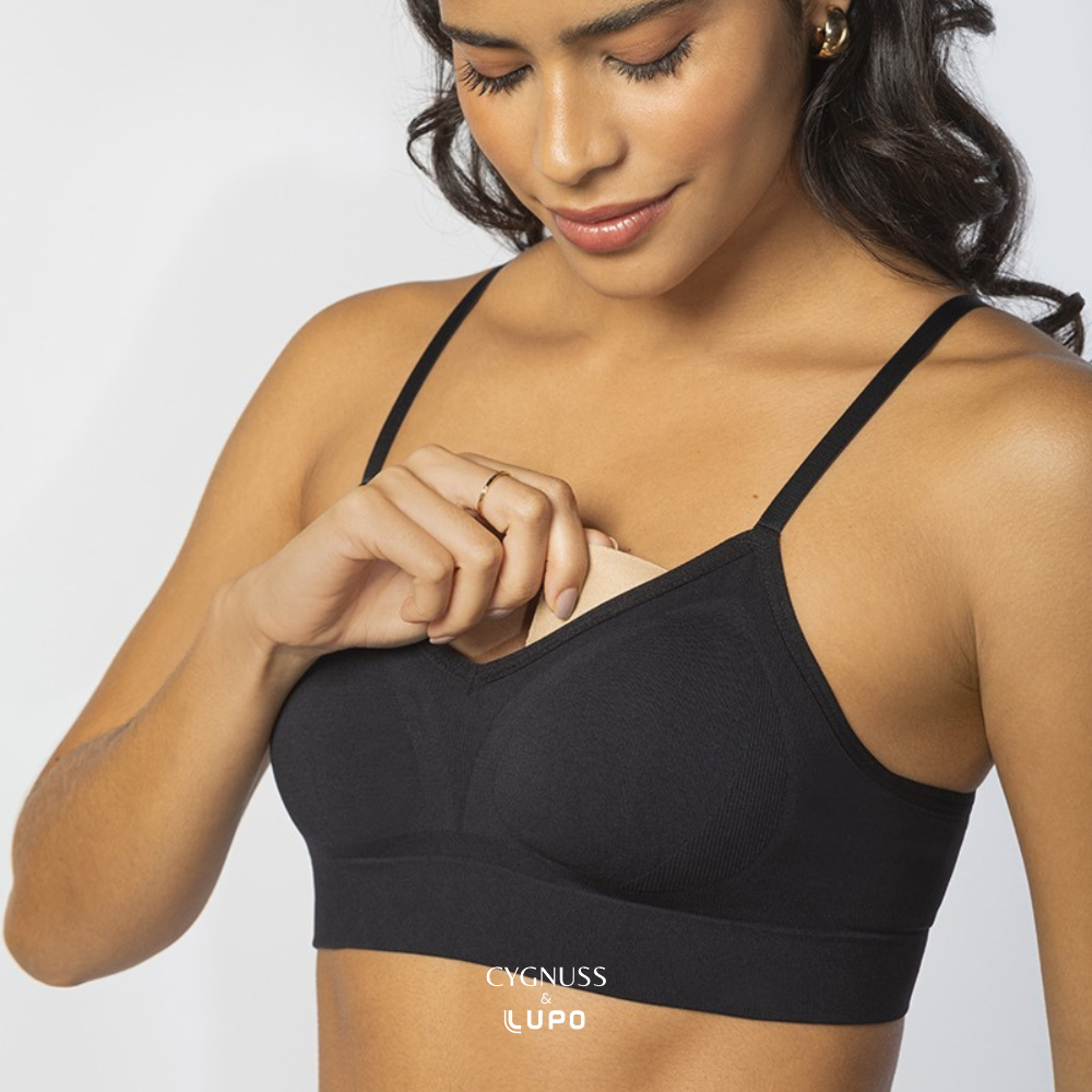 Mulher de pele morena e cabelos ondulados sorrindo enquanto ajusta a bojo removível de seu sutiã preto com alças finas. O modelo sem costuras oferece conforto, sustentação e respirabilidade. O logotipo da Cygnuss & Lupo está sobreposto na parte inferior da peça. Fundo branco minimalista.