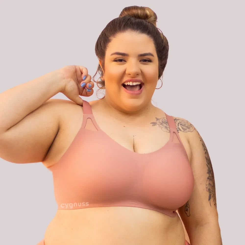 Mulher plus size sorridente vestindo o Sutiã Dreams na cor rosé, destacando o design sem costuras, as alças largas e os detalhes vazados próximos aos ombros. O tecido molda o corpo com conforto e sustentação. O logo da Cygnuss está visível na parte inferior do sutiã.