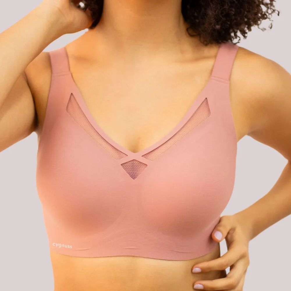 Mulher vestindo o Sutiã Dreams na cor rosé, destacando o design sem costuras, os detalhes vazados em tule no decote e as alças largas para melhor sustentação. A modelo posiciona as mãos na cintura, evidenciando o ajuste confortável do tecido. O logo da Cygnuss está visível na parte inferior do sutiã.