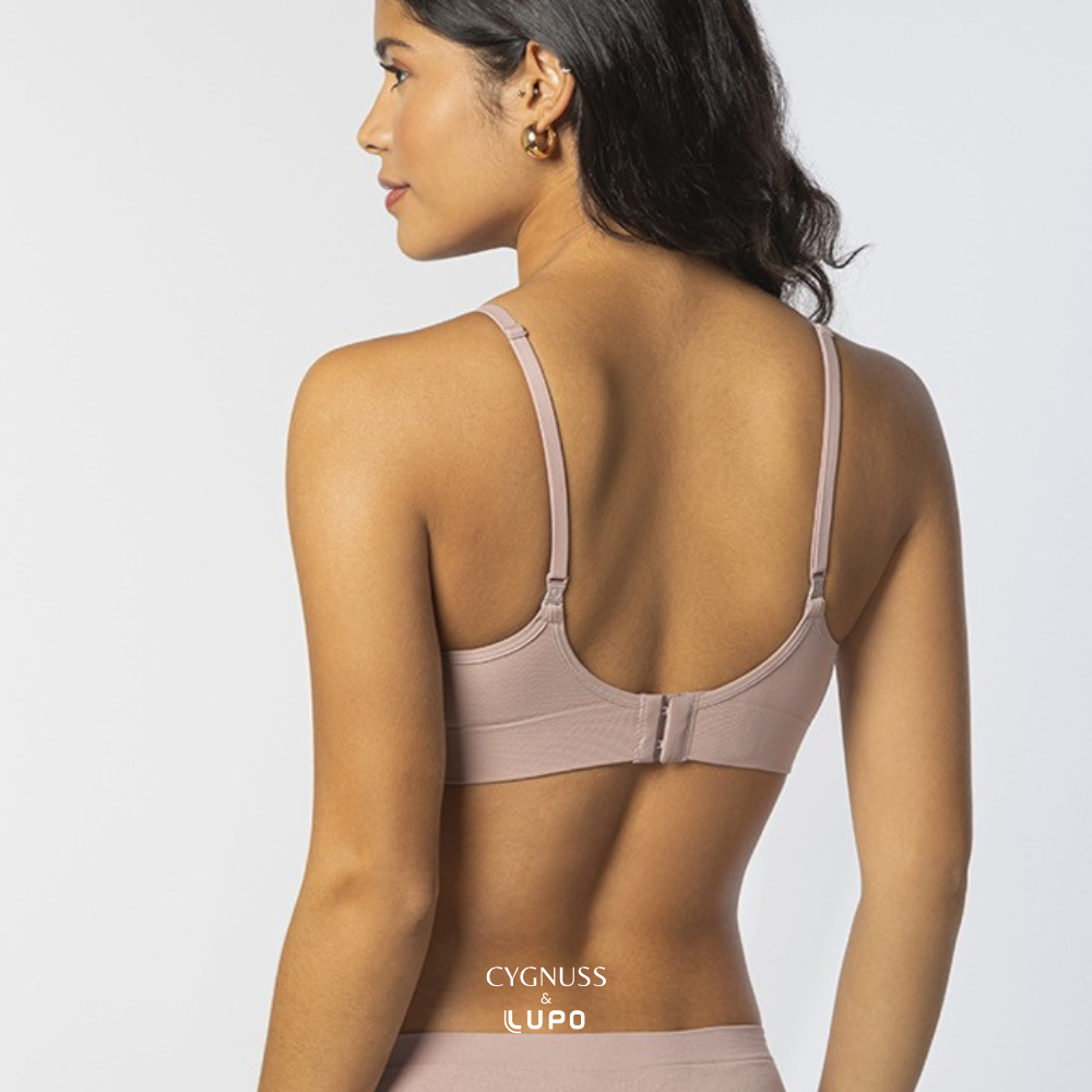 Vista traseira de uma mulher com pele morena e cabelos ondulados usando um sutiã rosa nude com alças finas ajustáveis. O design possui fecho traseiro e modelagem anatômica sem costuras, garantindo conforto e respirabilidade. O logotipo da Cygnuss & Lupo está sobreposto na parte inferior da peça. Fundo branco minimalista.