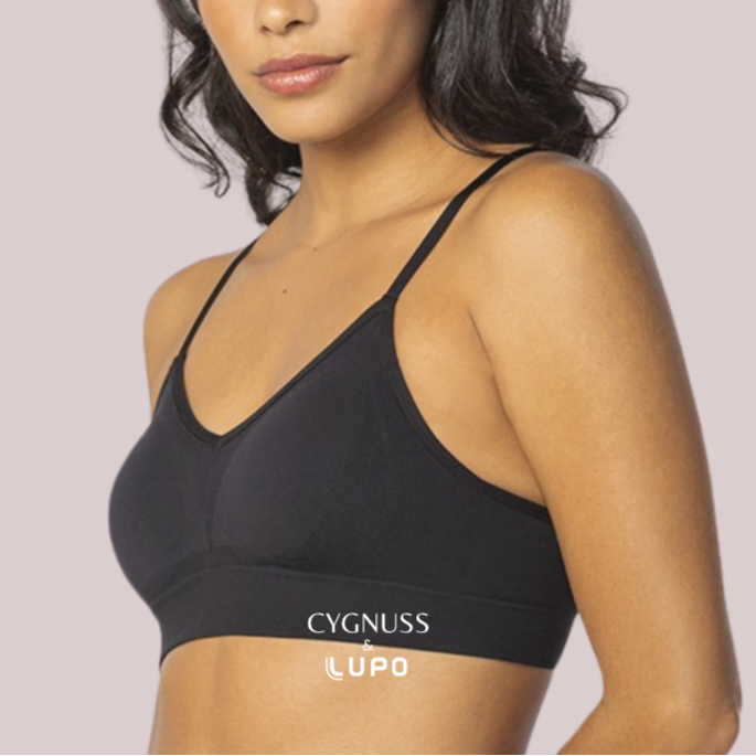 Mulher de pele morena e cabelos ondulados usando um sutiã preto com alças finas, sem costuras e com modelagem ajustada ao corpo. O tecido parece macio e confortável, oferecendo sustentação sem aro. O logotipo da Cygnuss & Lupo está sobreposto na parte inferior da peça. Fundo em tom nude rosado.
