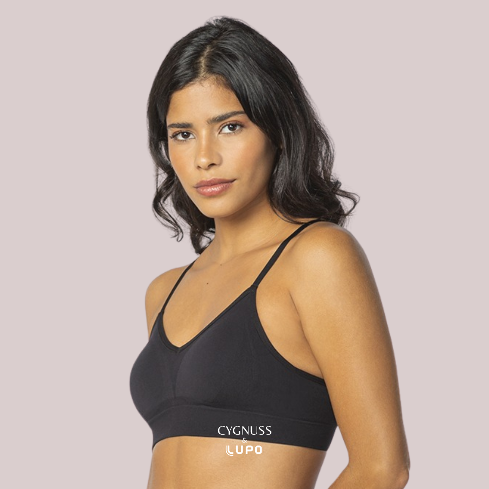 Mulher de pele morena e cabelos ondulados usando um sutiã preto sem costuras, com alças finas e modelagem anatômica. O tecido respirável e macio proporciona conforto e ajuste perfeito ao corpo. O logotipo da Cygnuss & Lupo está sobreposto na parte inferior da peça. Fundo nude rosado.