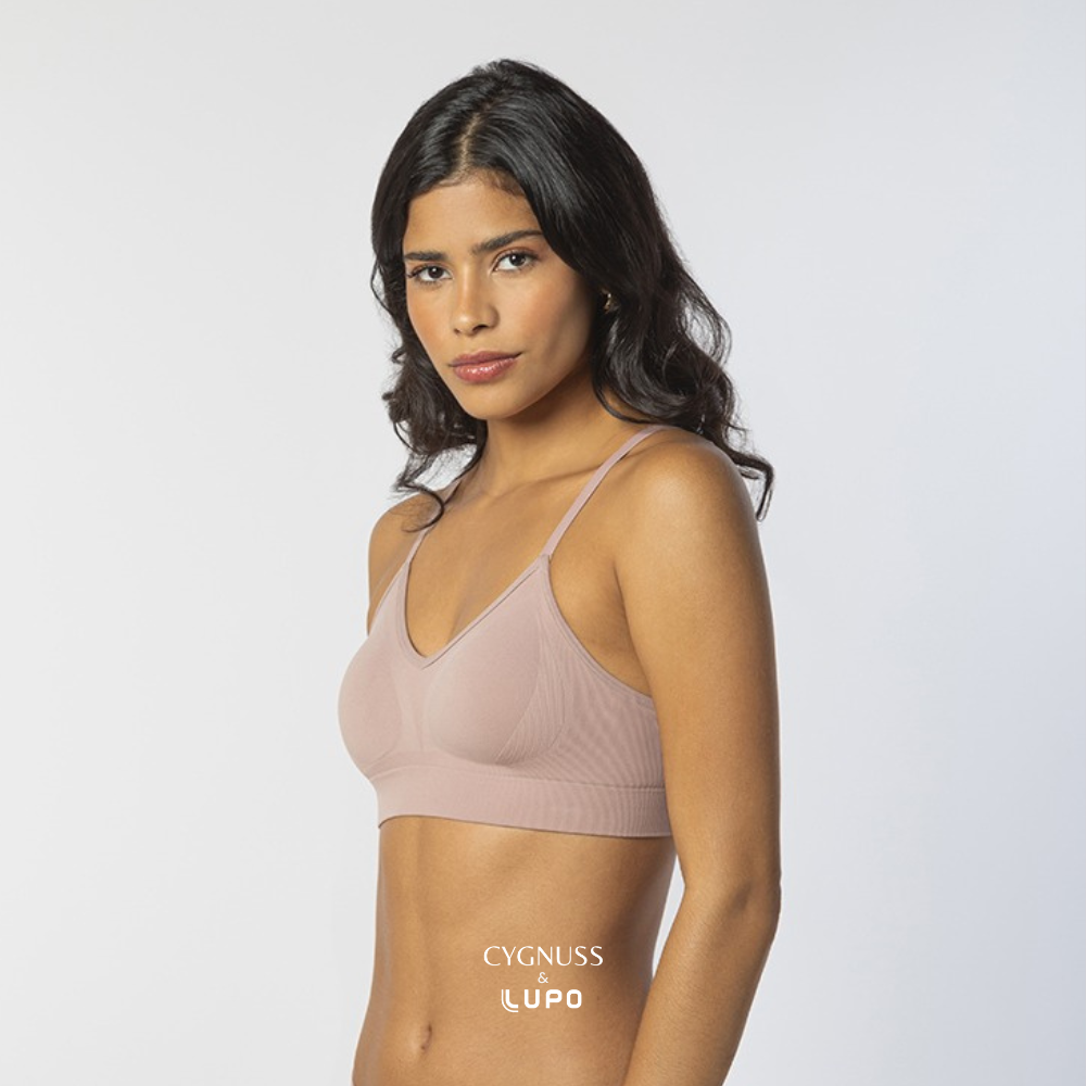 Mulher de pele morena e cabelos ondulados usando um sutiã rosa nude sem costuras, com alças finas e modelagem anatômica. O tecido leve e respirável proporciona conforto para uso o dia todo. O logotipo da Cygnuss & Lupo está sobreposto na parte inferior da peça. Fundo branco minimalista.