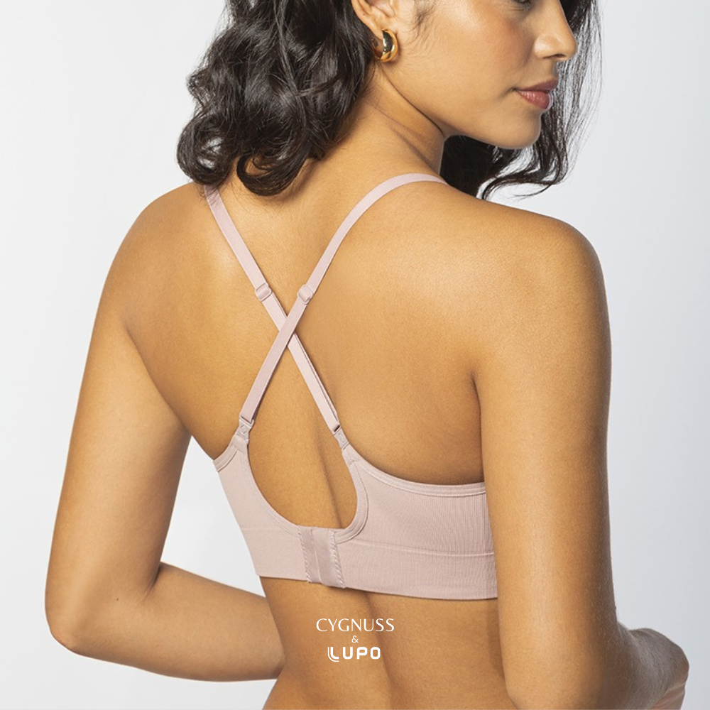 Vista traseira de uma mulher com pele morena e cabelos ondulados usando um sutiã rosa nude com alças finas cruzadas nas costas. O modelo possui fecho traseiro e modelagem anatômica sem costuras, garantindo conforto, respirabilidade e suporte natural. O logotipo da Cygnuss & Lupo está sobreposto na parte inferior da peça. Fundo branco minimalista.