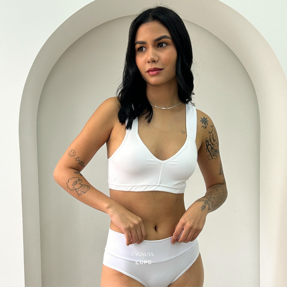 Mulher vestindo o conjunto Cygnuss composto pelo Sutiã Daily Original e a Calcinha Model na cor branca. O design sem costuras e as alças largas garantem conforto e sustentação. A modelo posa diante de um fundo minimalista com um arco decorativo, destacando a leveza e a sofisticação das peças.