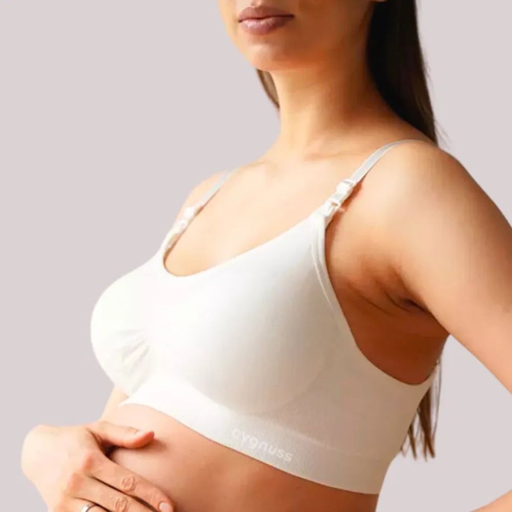 Mulher gestante usando o Sutiã Amamentação sem Bojo Cygnuss x Lupo na cor off-white. A imagem mostra a peça em ângulo lateral, com alças finas e clipe funcional para amamentação. A modelo apoia as mãos na barriga, transmitindo suavidade e acolhimento.