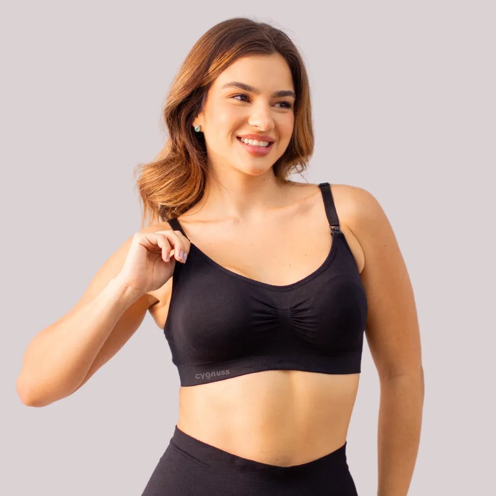 Mulher sorridente vestindo o Sutiã Amamentação sem Bojo na cor preta da collab Cygnuss x Lupo. A modelo está demonstrando a funcionalidade do clipe de abertura na alça, ideal para amamentação. O sutiã tem tecido canelado, ajuste confortável e leve franzido no centro do busto.