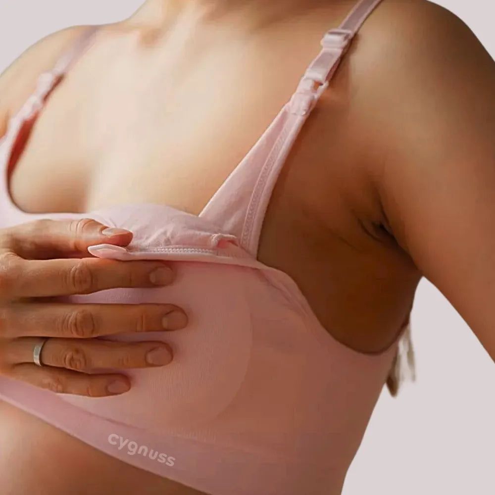 Close lateral de uma mulher usando o Sutiã Amamentação sem Bojo rosa claro da collab Cygnuss x Lupo. A imagem mostra o sutiã com a parte frontal aberta, revelando o espaço interno para amamentação. A modelo segura delicadamente o busto, destacando o conforto e a praticidade da peça.
