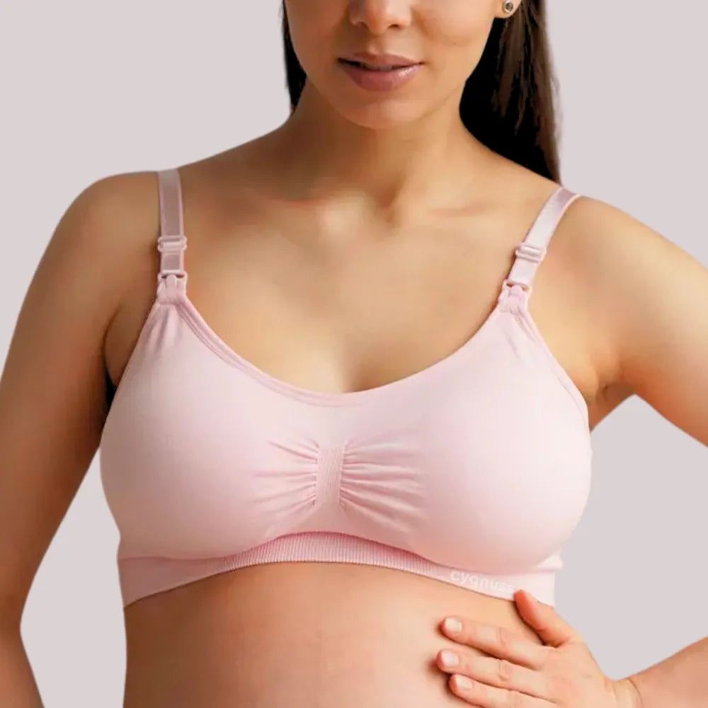 Mulher gestante vestindo o Sutiã Amamentação sem Bojo na cor rosa claro da collab Cygnuss x Lupo. O sutiã tem alças finas com abertura funcional para amamentação e franzido central no busto para melhor ajuste. A modelo está com uma das mãos apoiadas na barriga, destacando a proposta maternidade da peça.