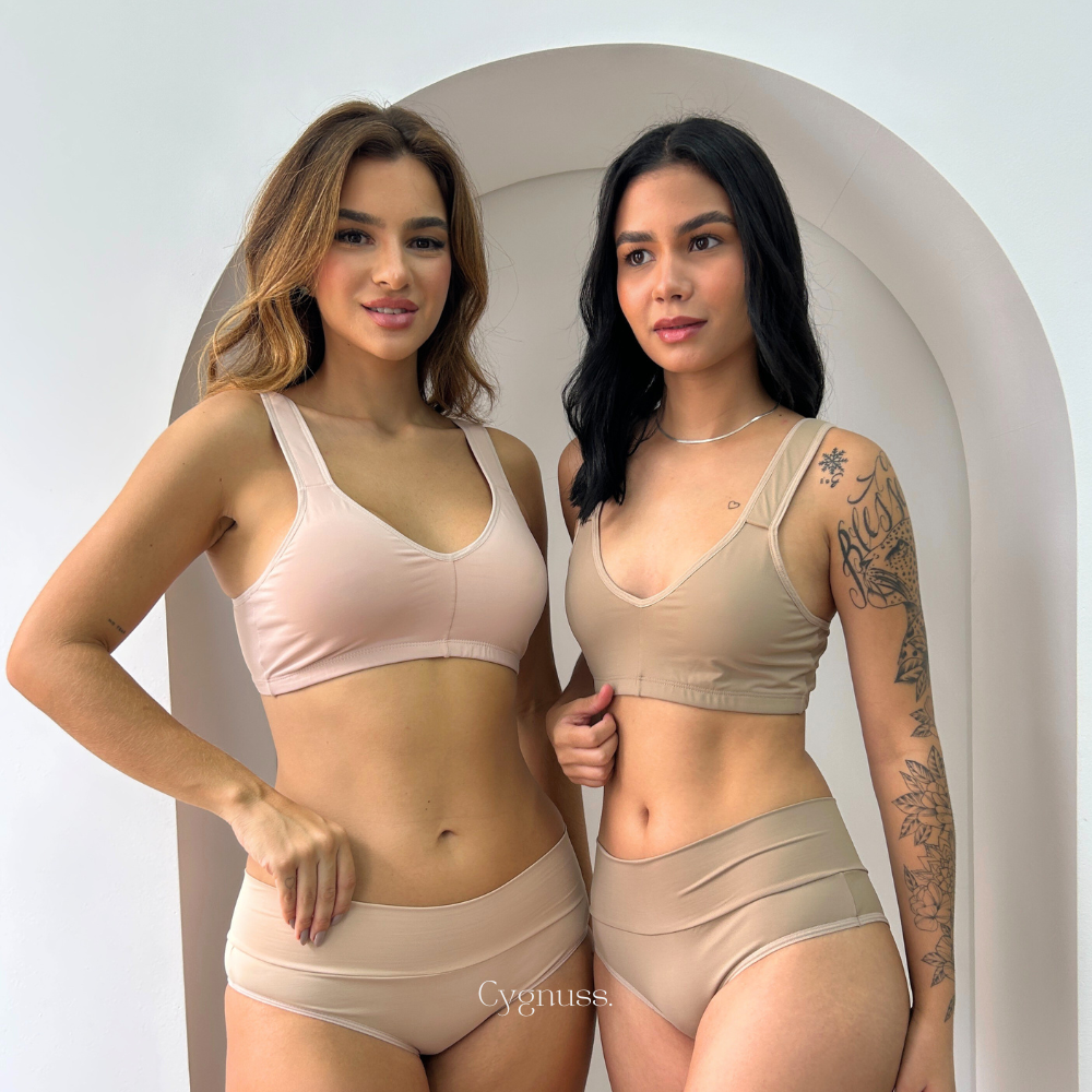 Duas mulheres vestindo o conjunto Cygnuss composto pelo Sutiã Daily Original e a Calcinha Model em tons de nude. O design sem costuras e as alças largas proporcionam conforto e sustentação. As modelos posam diante de um fundo minimalista com arco decorativo, destacando a sofisticação e a versatilidade das peças.