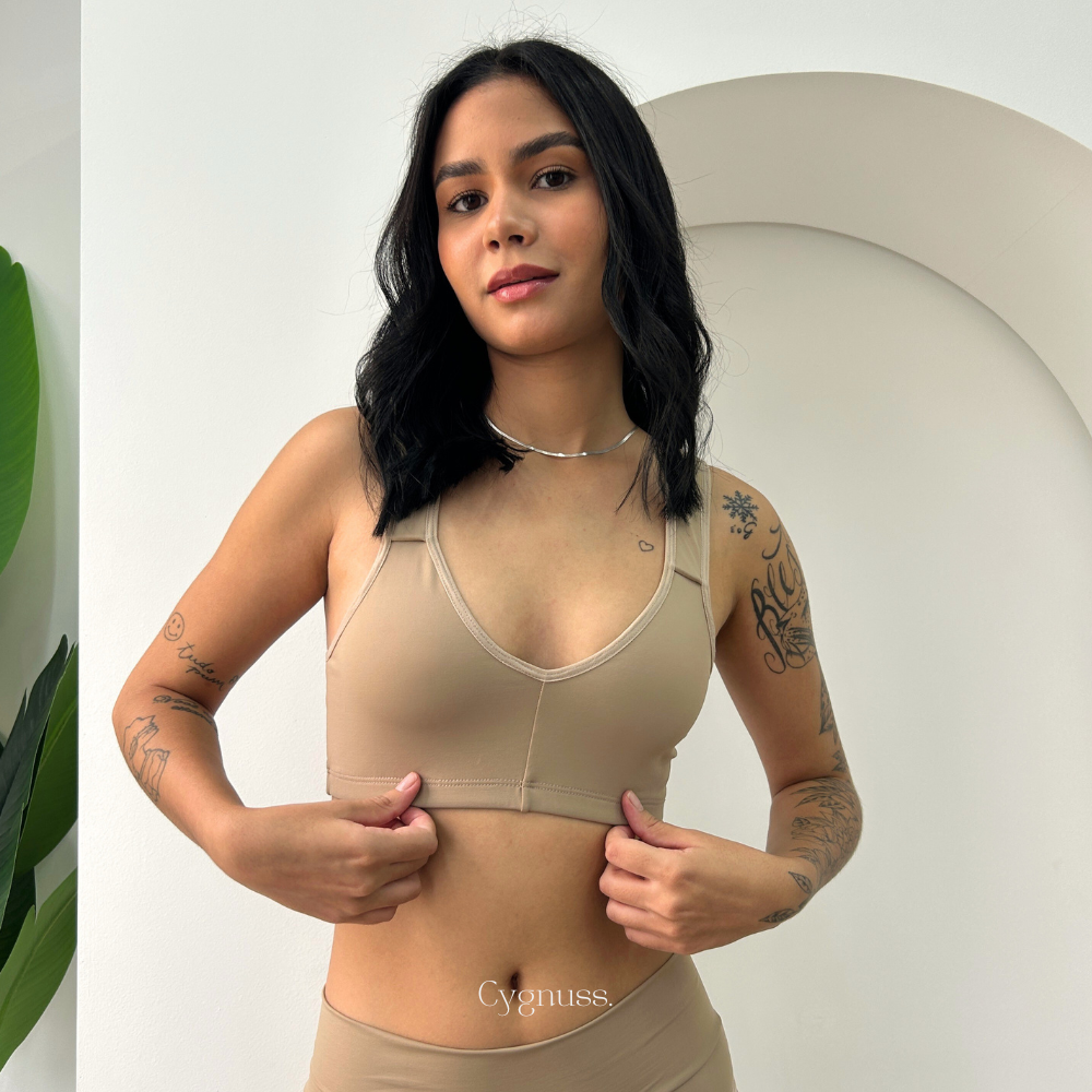 Mulher vestindo o Sutiã Daily Original na cor nude da Cygnuss. A imagem destaca o ajuste perfeito da peça sem costuras, proporcionando conforto e sustentação. A modelo segura suavemente a barra inferior do sutiã, evidenciando a elasticidade do tecido. O fundo minimalista com arco decorativo realça a sofisticação do produto.