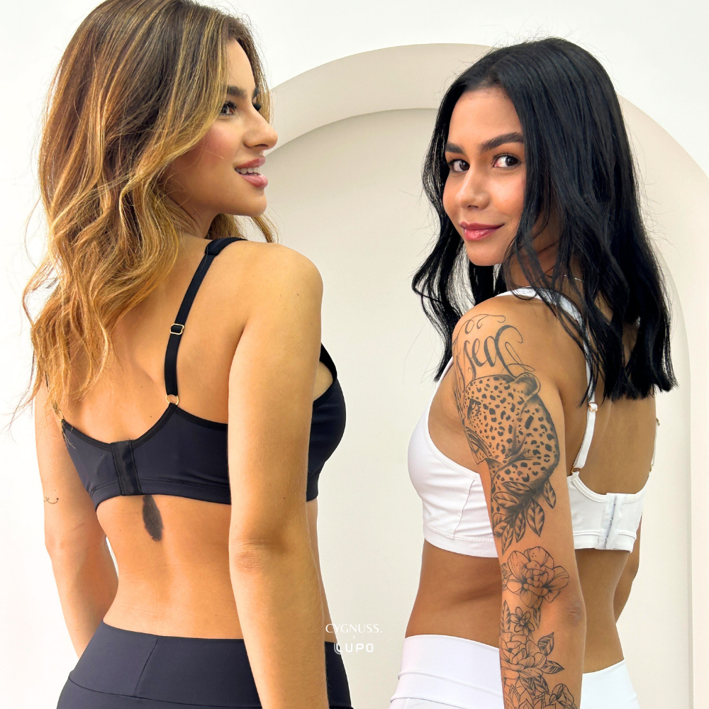 Duas mulheres mostrando a parte traseira do Sutiã Daily Original da Cygnuss, uma na cor preta e outra na cor branca. A imagem destaca o ajuste das alças reguláveis e o fecho traseiro, proporcionando sustentação e conforto. As modelos sorriem levemente, exibindo a elegância e praticidade do design. O fundo minimalista com arco decorativo complementa a composição.