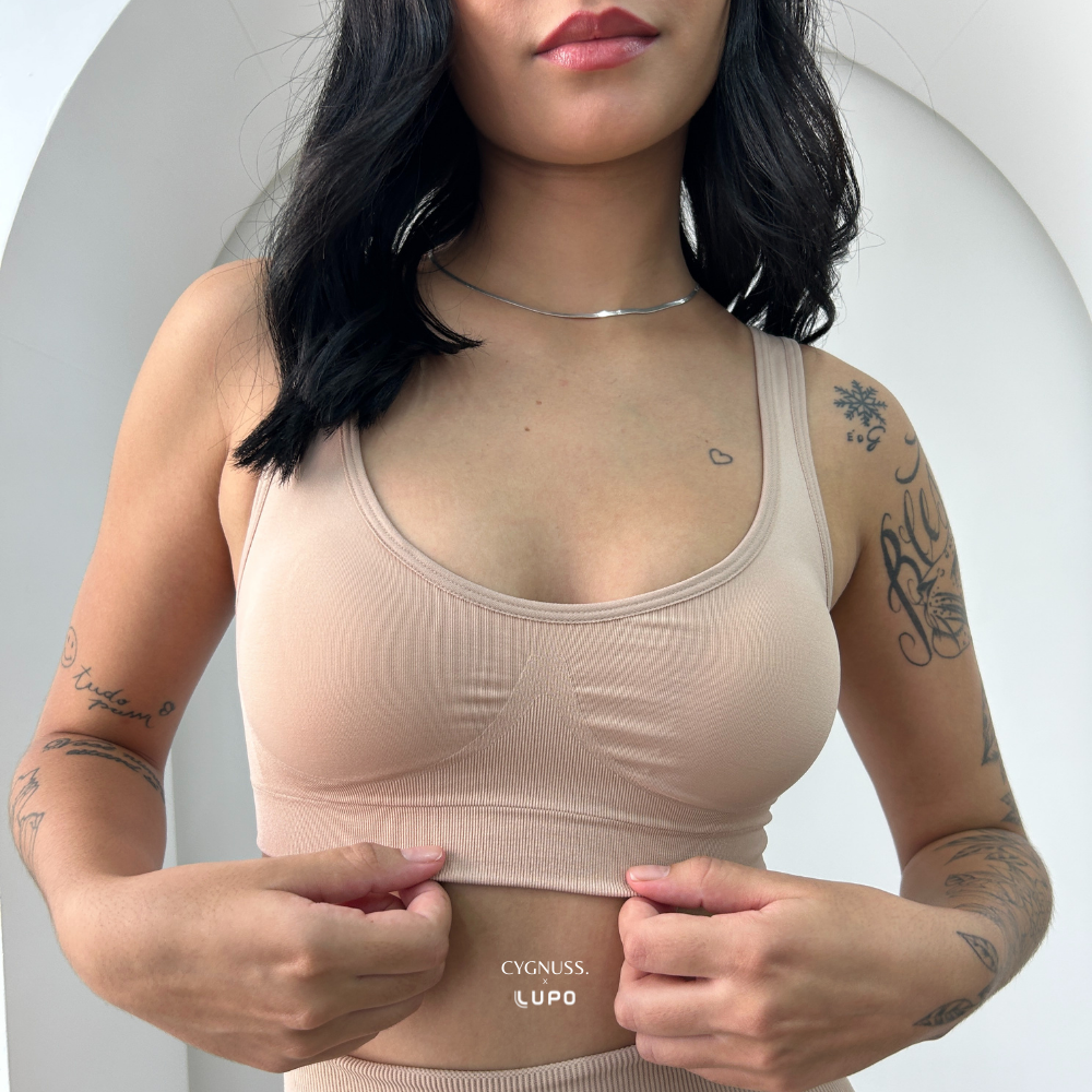Mulher com cabelos escuros usando top nude sem costura da Cygnuss em parceria com Lupo. A peça possui alças largas, bojo anatômico e cós reforçado. A modelo segura levemente a barra do top, evidenciando a elasticidade e o ajuste ao corpo. Fundo com arco branco moderno.