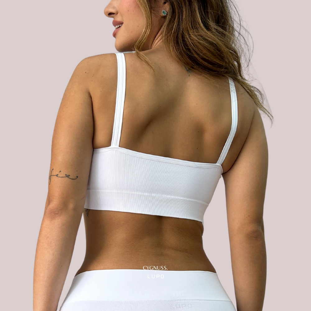 Mulher vestindo o Top Power na cor branca da Cygnuss, destacando a parte traseira da peça. O modelo sem costuras possui alças finas e um elástico reforçado abaixo do busto, garantindo conforto e sustentação. A textura canelada do tecido realça o design moderno e versátil.
