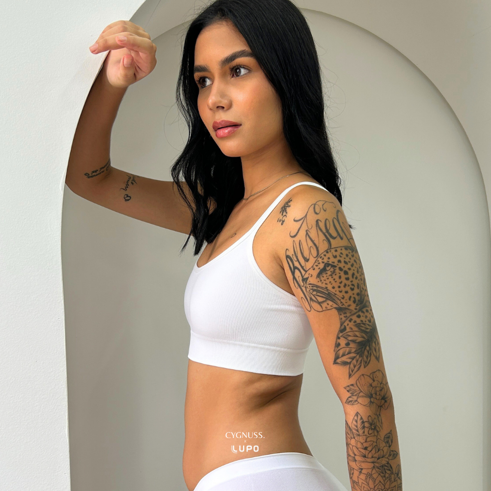 Mulher vestindo o Top Power na cor branca da Cygnuss, posando de perfil. A peça sem costuras apresenta alças finas e elástico reforçado abaixo do busto, garantindo conforto e sustentação. A textura canelada do tecido adiciona um toque moderno ao design. A modelo posa com uma expressão confiante diante de um fundo minimalista com arco decorativo