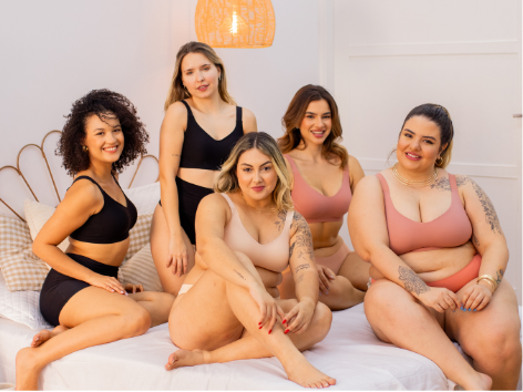 Grupo de cinco mulheres diversas usando lingeries confortáveis em tons neutros, sentadas em cama, representando autoestima, inclusão e bem-estar feminino.