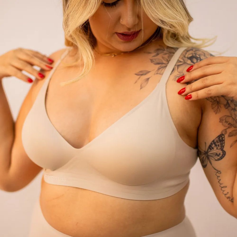 Mulher loira com unhas vermelhas vestindo o Sutiã Alça Fina Cygnuss na cor nude, ajustando as alças finas para um encaixe perfeito.