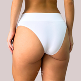 Vista traseira de uma mulher usando calcinha modeladora branca da marca Lupo em parceria com a Cygnuss. A peça tem cós alto, acabamento sem costura e modelagem anatômica. Fundo lilás claro.