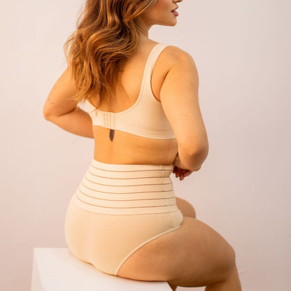 Imagem de mulher usando a Calcinha Modeladora Model - Seca Barriga, vista de costas. A peça de alta compressão é de tom bege, com listras horizontais na faixa superior, proporcionando controle da barriga e modelagem eficiente da silhueta.