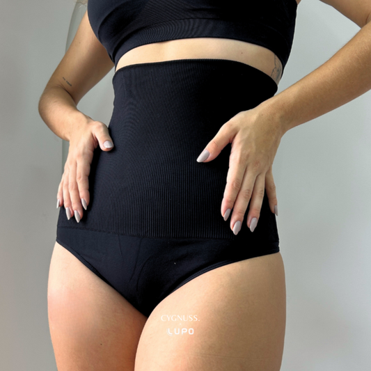 Mulher usando uma calcinha modeladora preta de cintura alta com efeito de compressão suave. O tecido canelado sem costuras proporciona ajuste confortável e modelagem definida, realçando a silhueta. A modelo posiciona as mãos na cintura, destacando o efeito modelador da peça. O logotipo da Cygnuss & Lupo está sobreposto na parte inferior da imagem.