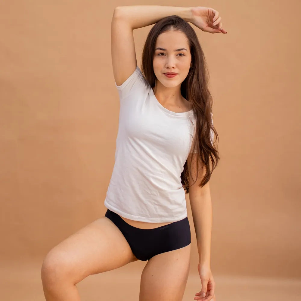 Mulher usando a Calcinha Soft preta da Cygnuss e camiseta branca, em pose descontraída com um braço levantado, contra fundo bege, evidenciando o caimento leve e o conforto da peça no corpo.