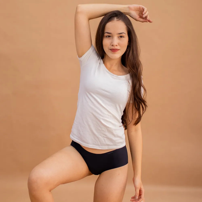 Mulher usando a Calcinha Soft preta da Cygnuss e camiseta branca, em pose descontraída com um braço levantado, contra fundo bege, evidenciando o caimento leve e o conforto da peça no corpo.