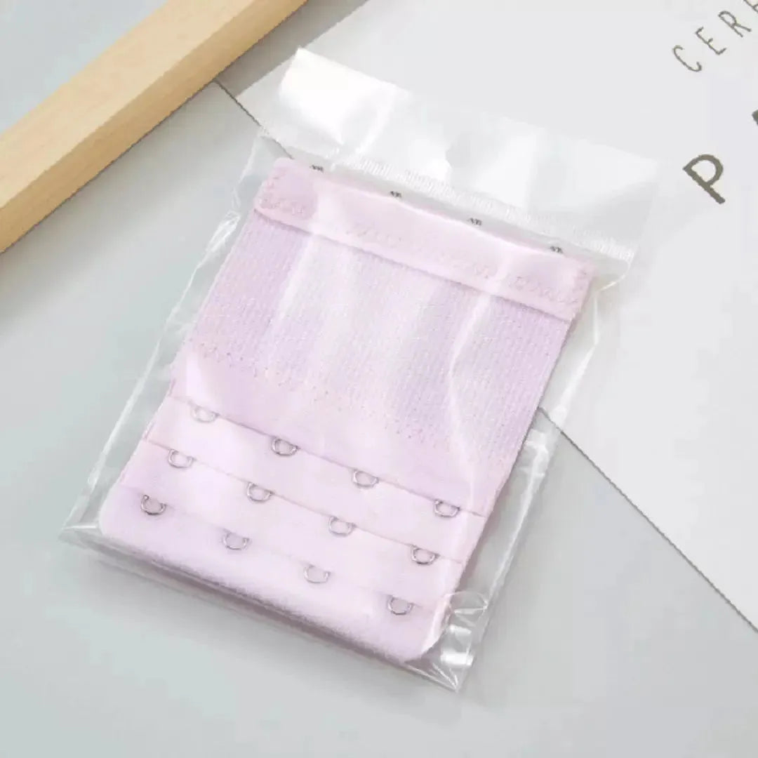 Extensor de sutia lilas com tres niveis de ajuste e ganchos metalicos, embalado individualmente em plastico transparente. Ideal para proporcionar mais conforto e flexibilidade ao vestir o sutia. Fundo claro e minimalista.