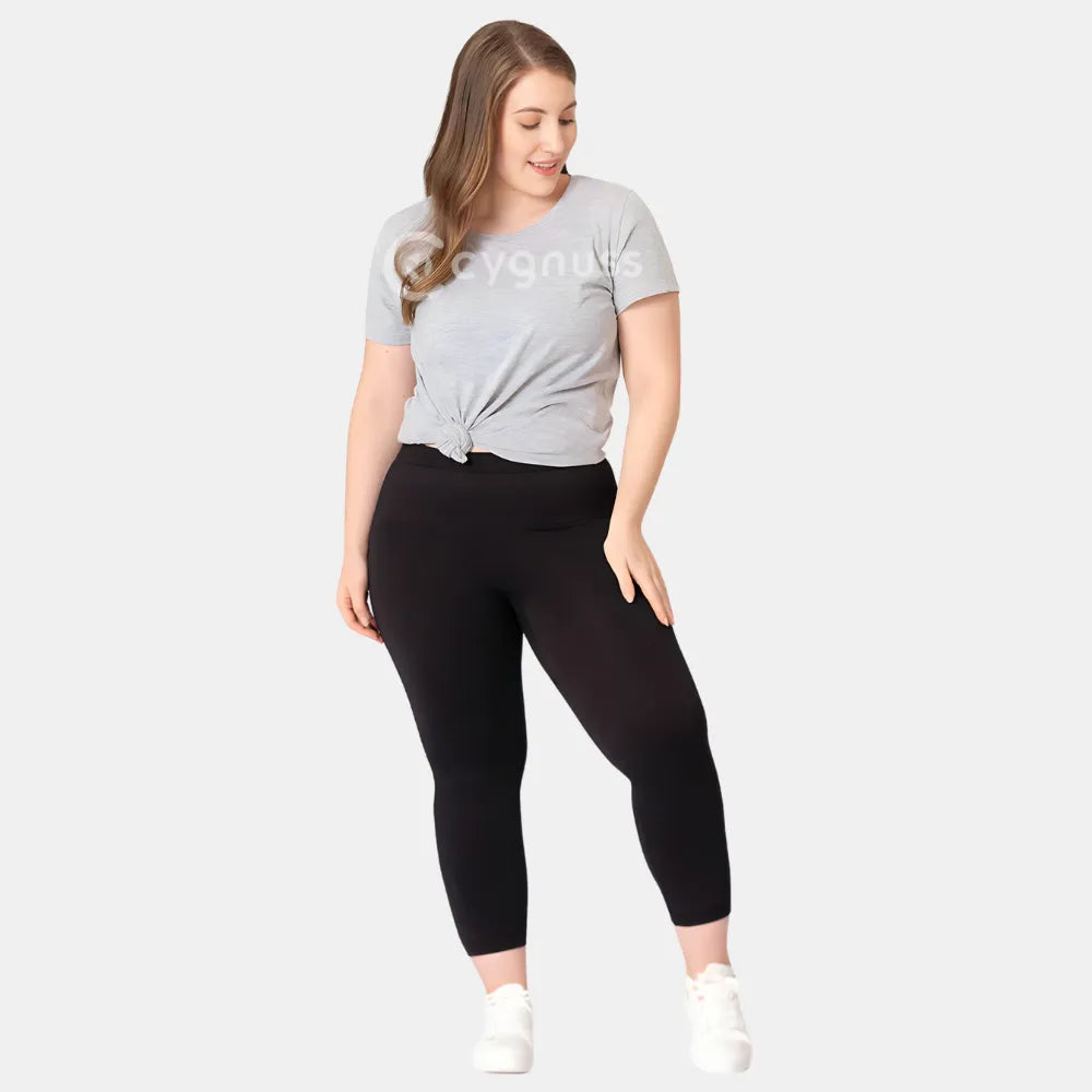 Mulher plus size usando a Legging Jade Fit Modeladora de Cós Alto na cor preta, combinada com camiseta cinza amarrada e tênis branco, em pose descontraída que evidencia o conforto e a versatilidade da peça no dia a dia.

