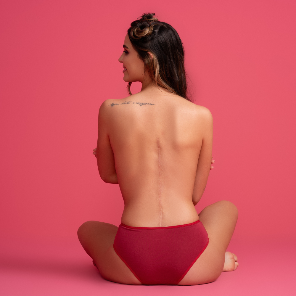 Modelo de calcinha vermelha vista de costas. A peça proporciona conforto e estilo, destacando a cor vibrante e o design elegante em um fundo rosa vibrante.