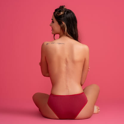 Modelo de calcinha vermelha vista de costas. A peça proporciona conforto e estilo, destacando a cor vibrante e o design elegante em um fundo rosa vibrante.