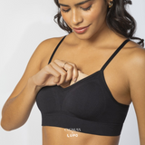 Mulher de pele morena e cabelos ondulados sorrindo enquanto ajusta a bojo removível de seu sutiã preto com alças finas. O modelo sem costuras oferece conforto, sustentação e respirabilidade. O logotipo da Cygnuss & Lupo está sobreposto na parte inferior da peça. Fundo branco minimalista.