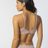 Vista traseira de uma mulher com pele morena e cabelos ondulados usando um sutiã rosa nude com alças finas ajustáveis. O design possui fecho traseiro e modelagem anatômica sem costuras, garantindo conforto e respirabilidade. O logotipo da Cygnuss & Lupo está sobreposto na parte inferior da peça. Fundo branco minimalista.