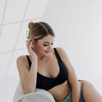 Mulher sentada em um sofá claro, com expressão suave e mão apoiada no rosto, vestindo o Sutiã FIT preto. A peça possui alças finas, bojo removível e acabamento sem costuras, oferecendo elegância, conforto e leveza no visual.