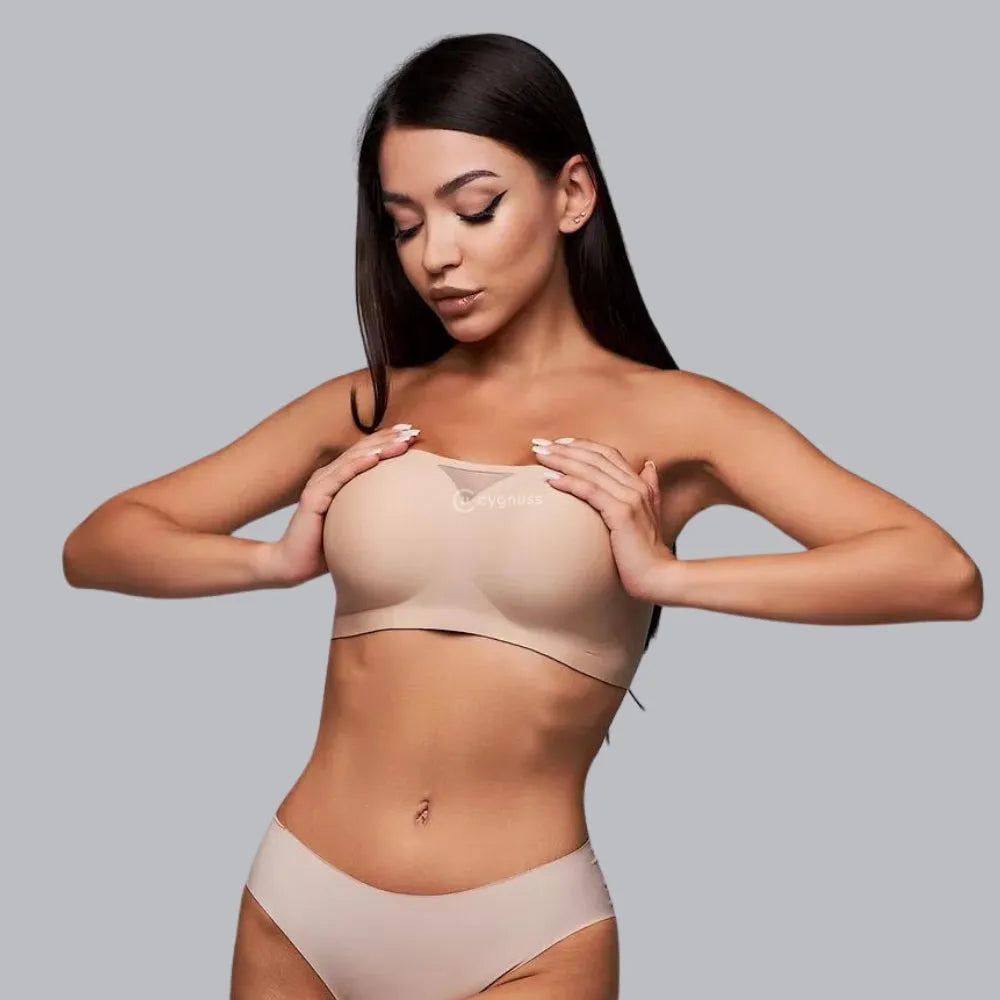 Modelo usando conjunto bege com sutiã tomara que caia Cygnuss, ajustando o busto com as mãos; peça sem costura com detalhe em tule no centro.